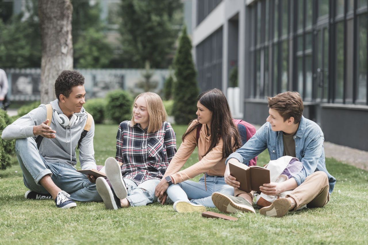smiling teenagers sitting on grass, talking and holding books